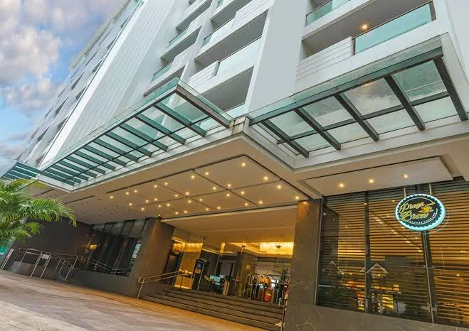 A modern hotel entrance with a glass canopy, well-lit facade, and large windows. There’s a neon sign near the entrance and luggage carts are visible inside the lobby area. The building has multiple floors.