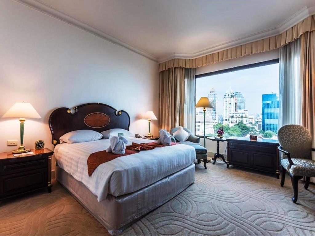 A hotel room with a large bed, two bedside tables with lamps, an armchair, a desk, and a large window offering a city view with tall buildings and greenery.