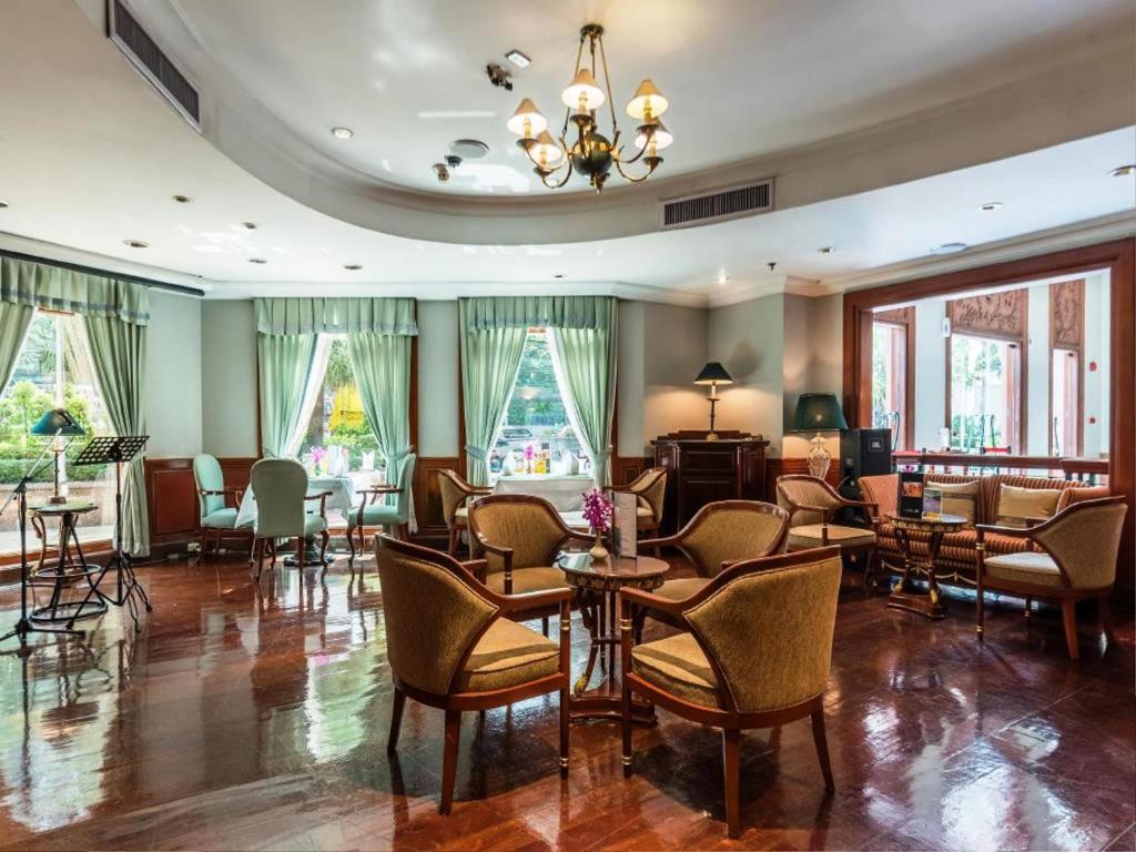 A cozy, elegant lounge with wooden floors, cushioned armchairs, small tables, and large windows with green curtains letting in natural light. A chandelier hangs from the ceiling, and plants are visible outside.