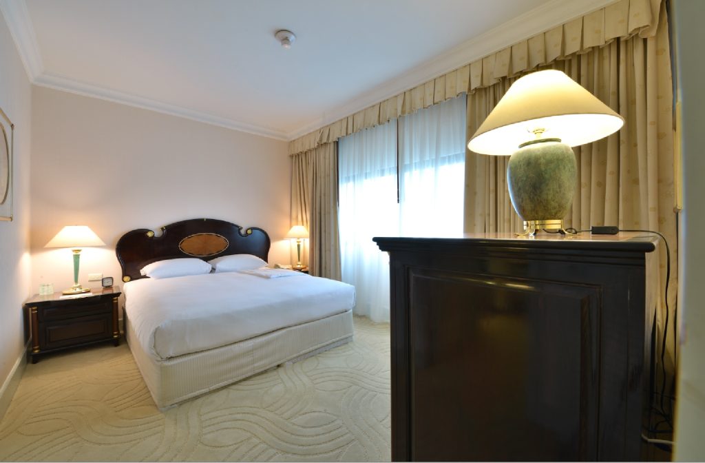 A neatly made double bed in a hotel room with two bedside lamps, a large window with curtains, light-colored walls, and a dresser with a lamp in the foreground. The room has a calm, inviting atmosphere.