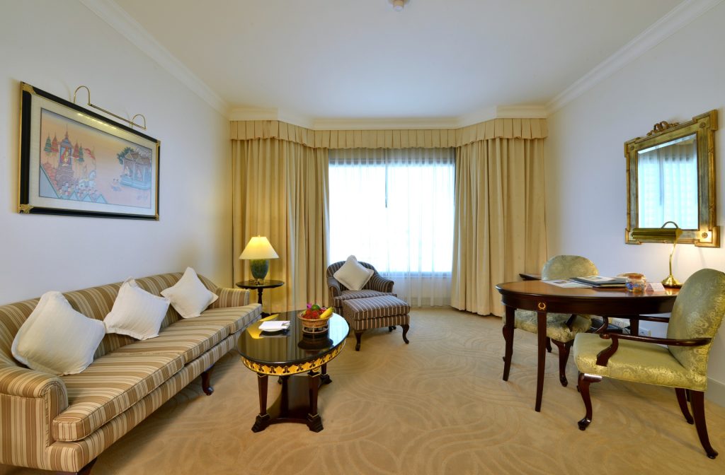 A well-lit hotel room with a striped sofa, armchair, and coffee table topped with fruit, a desk with two chairs, a large window with drapes, a lamp, and framed artwork on the walls.
