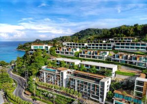 Aerial view of a modern hillside resort with multiple white buildings and balconies, surrounded by lush greenery, overlooking a winding road and a turquoise sea under a partly cloudy sky.