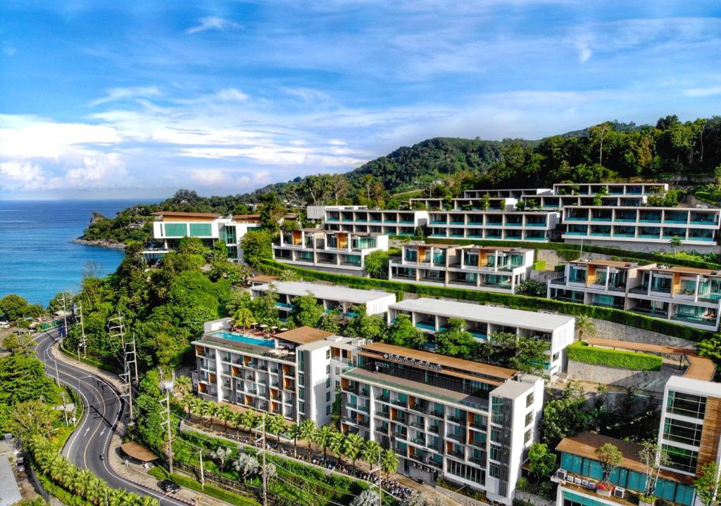 Aerial view of a modern hillside resort with multiple white buildings and balconies, surrounded by lush greenery, overlooking a winding road and a turquoise sea under a partly cloudy sky.