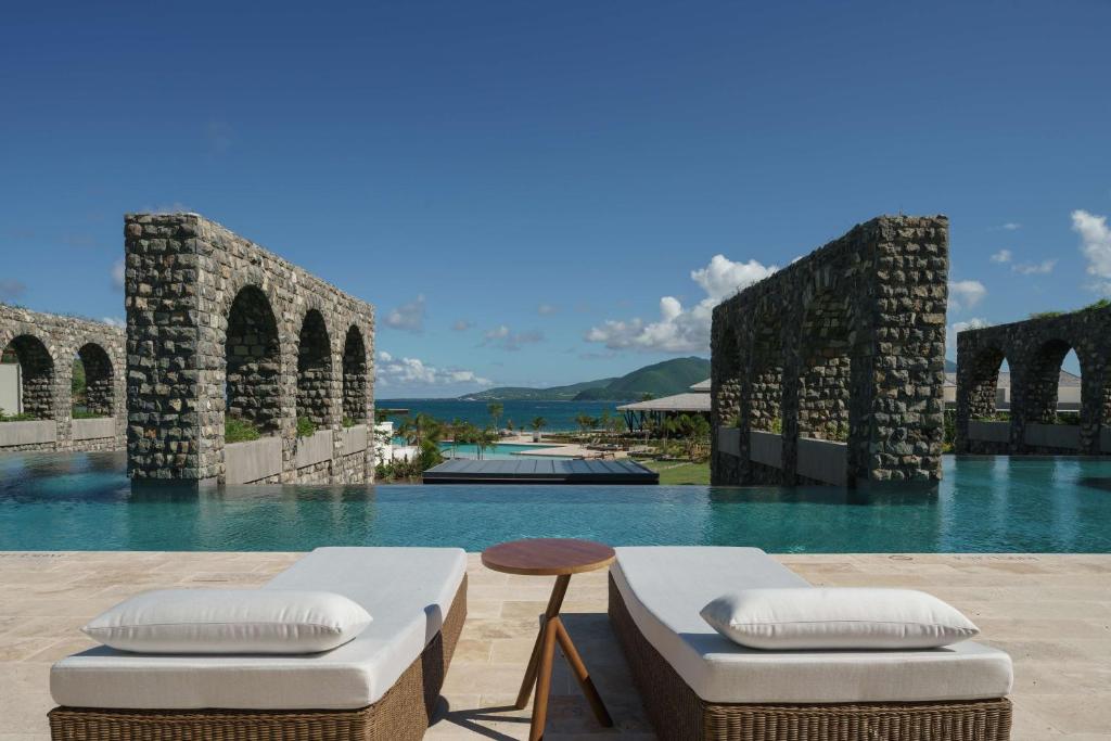 Two lounge chairs face a luxurious infinity pool bordered by tall stone arches, with a view of the ocean and distant green hills under a bright blue sky.