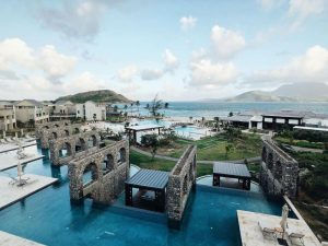 A luxury seaside resort features stone arches over blue swimming pools, surrounded by lush greenery, modern buildings, and overlooking mountains and the ocean under a partly cloudy sky.
