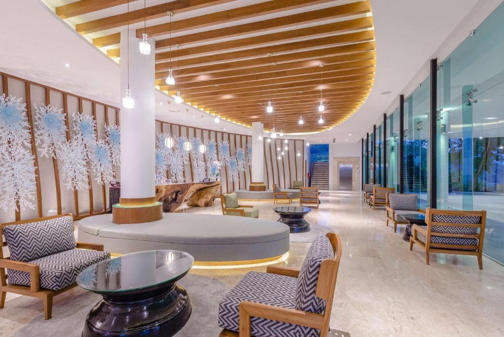 Modern hotel lobby with stylish wood accents on the ceiling, artistic wall decor, round sofas, patterned armchairs, glass tables, marble floors, and large floor-to-ceiling windows letting in natural light.