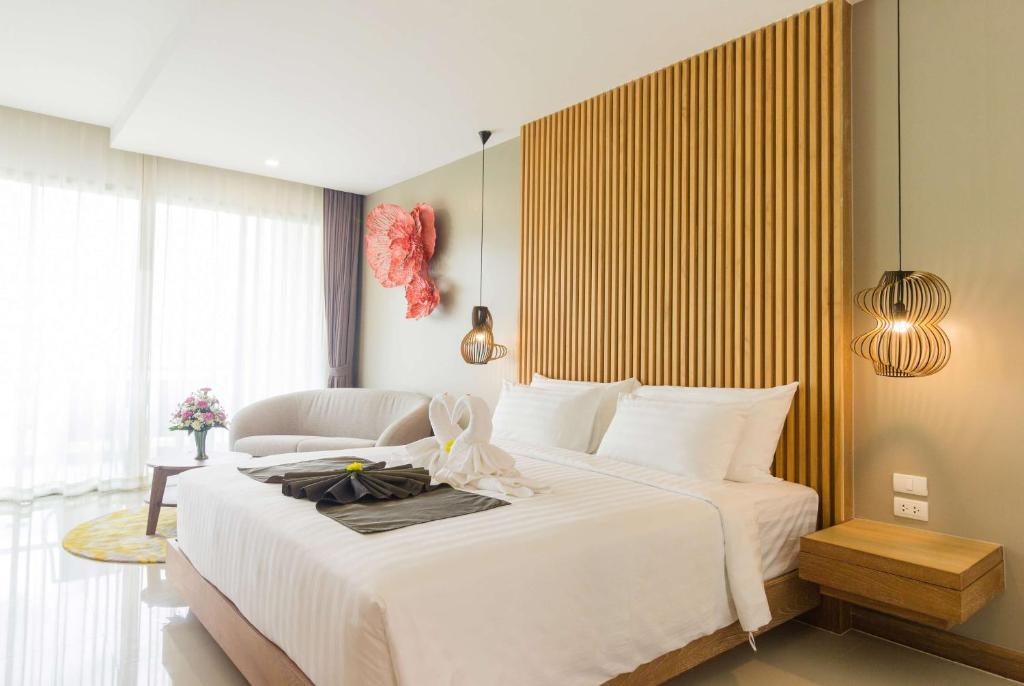 Bright, modern hotel room with a large bed, white linens, towel swans, a wooden accent wall, pendant lights, a sofa, round table with flowers, and large windows with sheer curtains letting in natural light.
