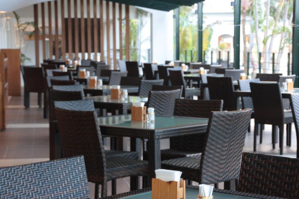 A modern indoor restaurant with empty dark wicker chairs and glass-top tables, each set with napkin holders and condiments. Large windows let in natural light, and plants are visible outside.