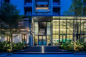 The illuminated exterior of Cross Vibe Bangkok Sukhumvit shows a modern glass-front entrance, lush green plants, steps leading up, and a tall sign with the hotels name at night.