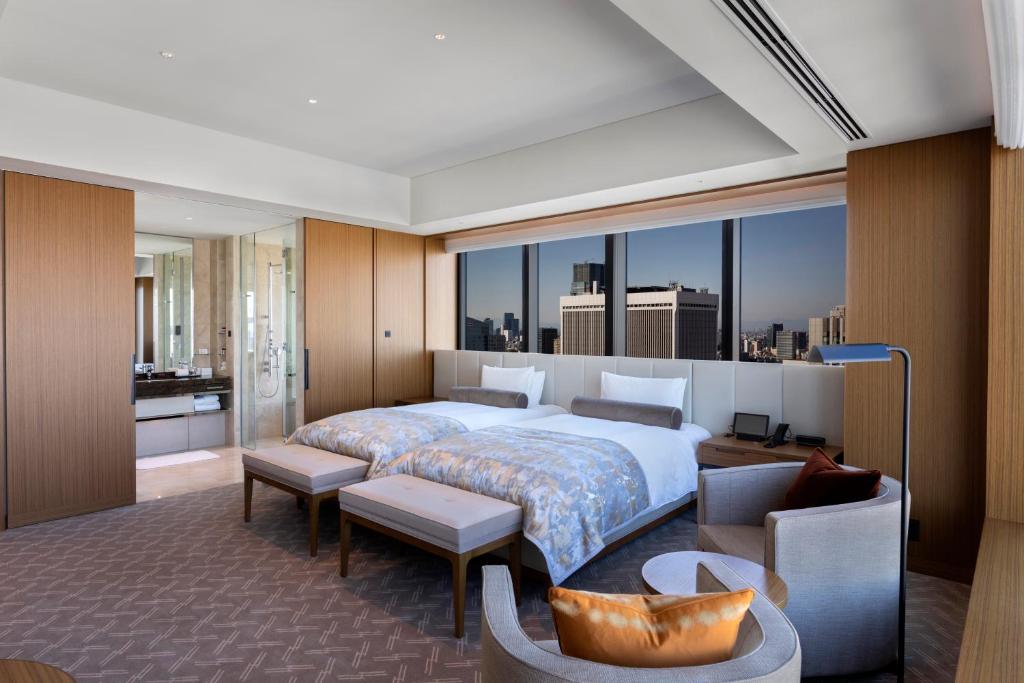 Modern hotel room with two double beds, large floor-to-ceiling windows offering a city view, a seating area with an armchair and table, and a bathroom visible through a glass wall. Decor features neutral tones with wood accents.