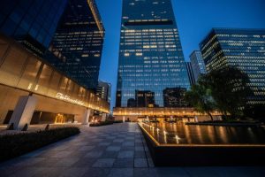 Modern cityscape at dusk featuring tall glass office buildings, illuminated windows, a reflective water feature, and the illuminated The Ambassador sign near the entrance of a building.