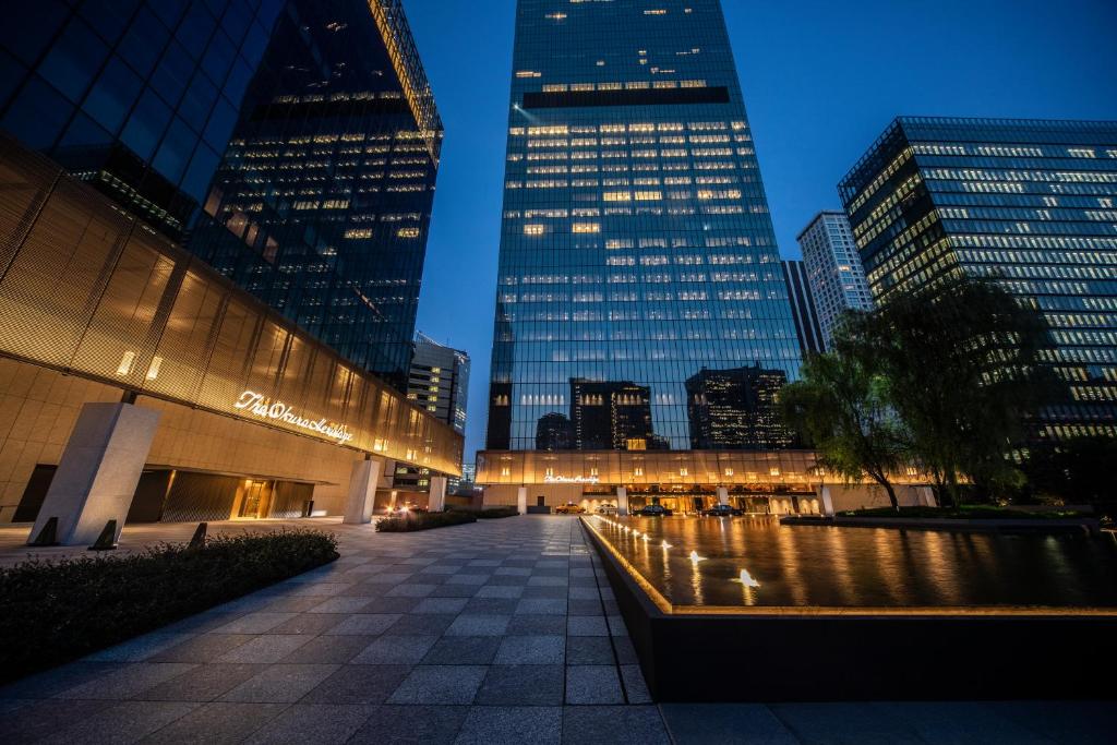 Modern cityscape at dusk featuring tall glass office buildings, illuminated windows, a reflective water feature, and the illuminated The Ambassador sign near the entrance of a building.