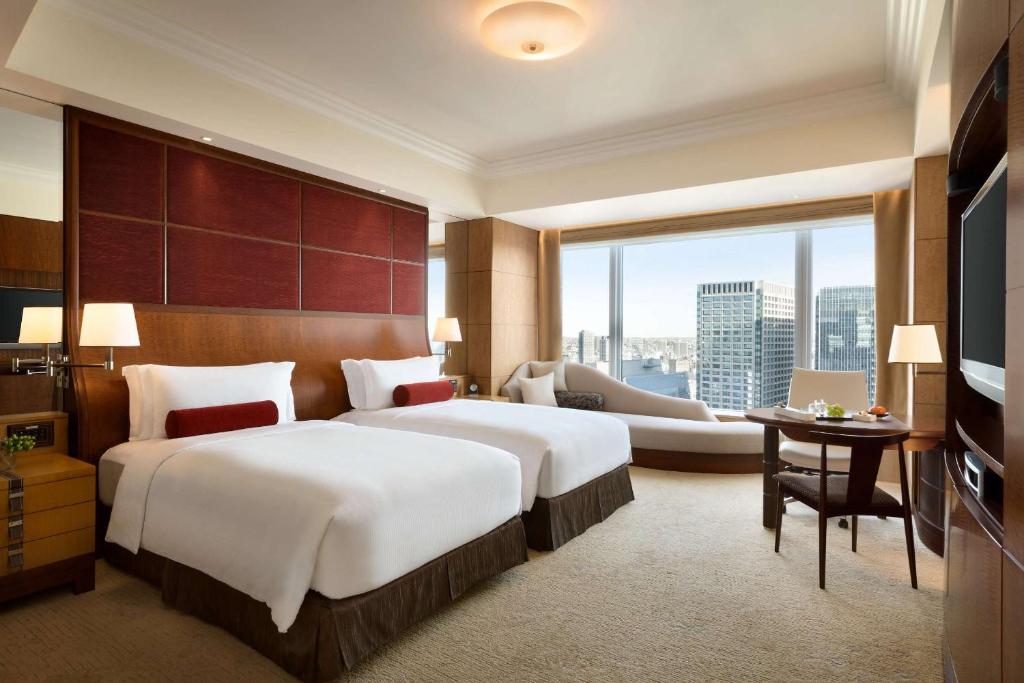 A hotel room with two double beds, white bedding, red accent pillows, a chaise lounge by a large window overlooking city buildings, a table with chairs, and warm-toned modern decor.