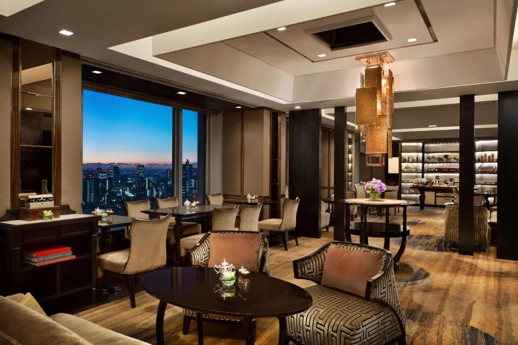 Elegant hotel lounge with plush chairs, tables, and soft lighting. Large windows reveal a city skyline at dusk. Shelves with books and decor line the walls, and fresh flowers are arranged throughout the space.