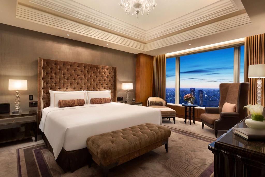 Luxurious hotel room with a large bed, tufted headboard, bench, armchairs, lamps, and a window offering a city view at dusk. Neutral tones and elegant decor create a comfortable, upscale atmosphere.