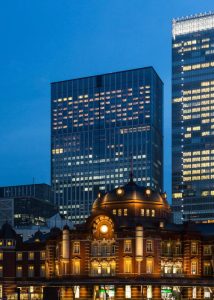 Historic Tokyo Station with its red brick facade and domed roof is illuminated at dusk, set against the backdrop of modern glass skyscrapers in the city’s Marunouchi district.
