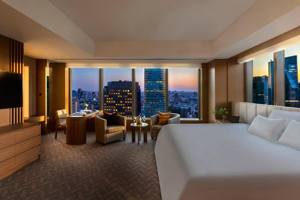 A modern hotel room with a large bed, seating area, and floor-to-ceiling windows overlooking a city skyline at sunset.