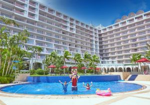 A man and three children play and pose happily in a round hotel pool, surrounded by palm trees, red umbrellas, and a large multi-story resort building under a blue sky.