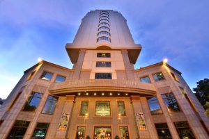 A tall, modern hotel building with a wide entrance, large windows, and the sign Evergreen Laurel Hotel above the doorway, photographed from a low angle at dusk under a blue sky.