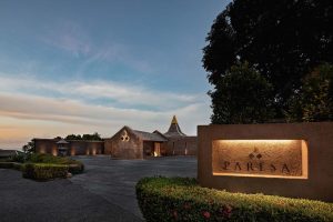 The image shows the entrance to a luxury resort at dusk, with warm lighting illuminating a sign that reads Paresa. The building has a modern design with stone walls and landscaped greenery around the driveway.