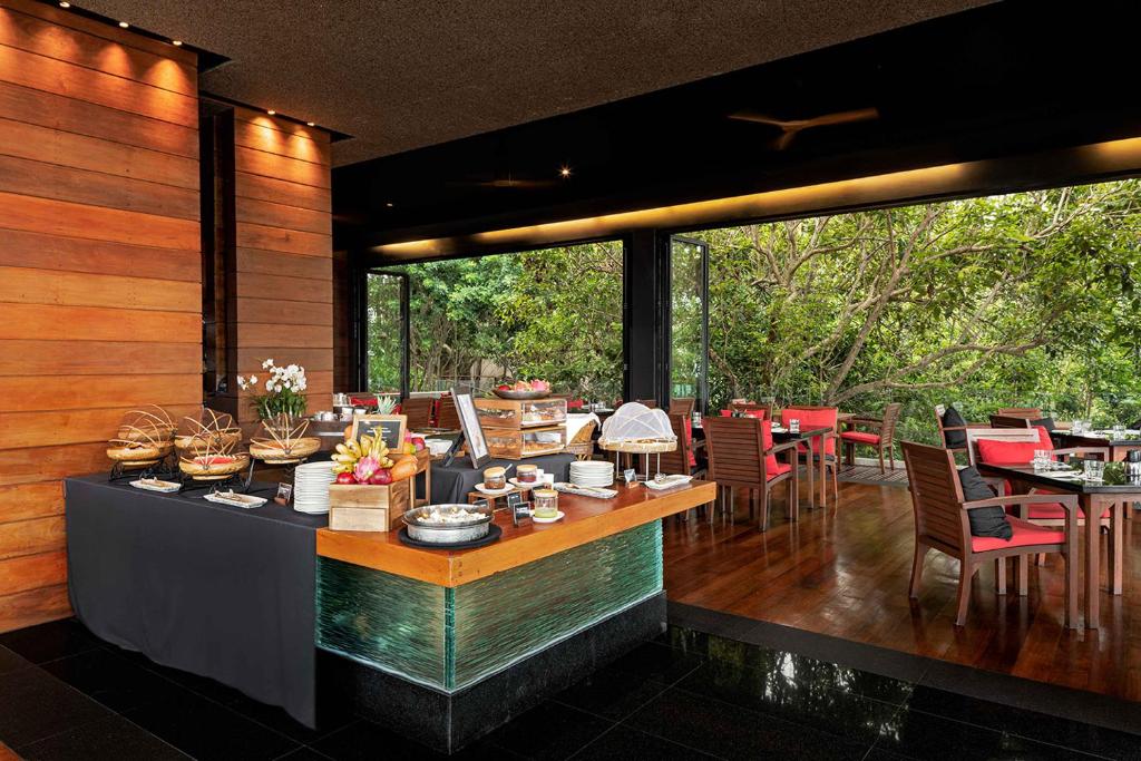 A modern restaurant with wooden walls and floor features a buffet table set with various breakfast items. Large windows reveal lush green trees outside, and red chairs are arranged around dining tables in the seating area.
