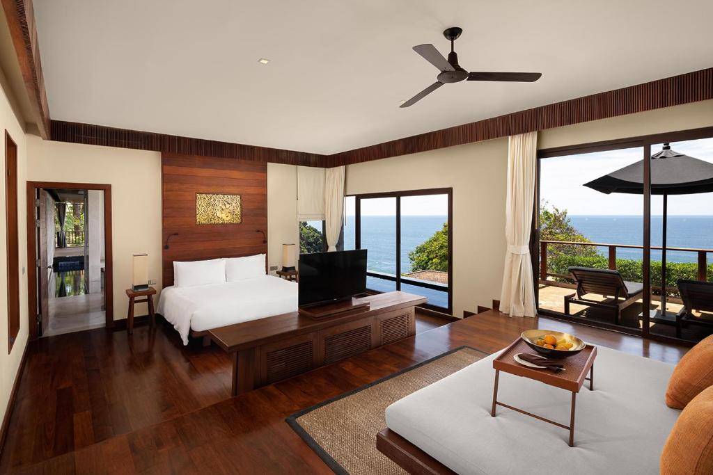 Spacious, modern hotel room with wooden floors, large bed, TV, sofa, and sliding glass doors opening to a balcony with ocean views, lounge chairs, and an umbrella. Natural light fills the elegant, airy space.