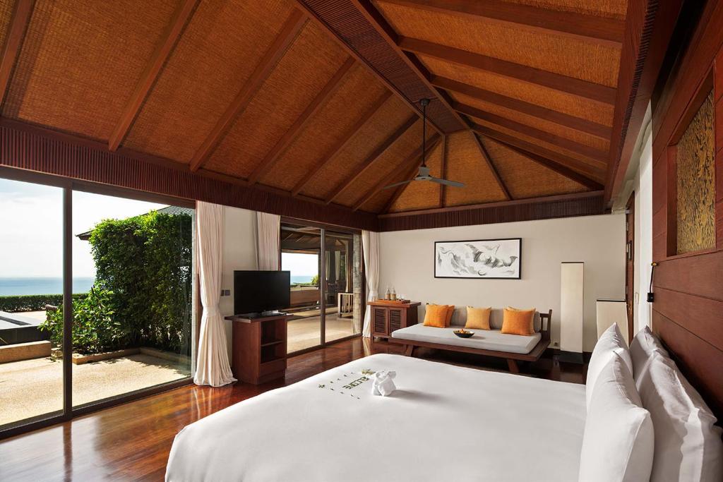 Spacious tropical villa bedroom with wooden floors, high vaulted ceiling, large bed, TV, sofa with orange cushions, and floor-to-ceiling windows opening to a patio with ocean views and greenery.
