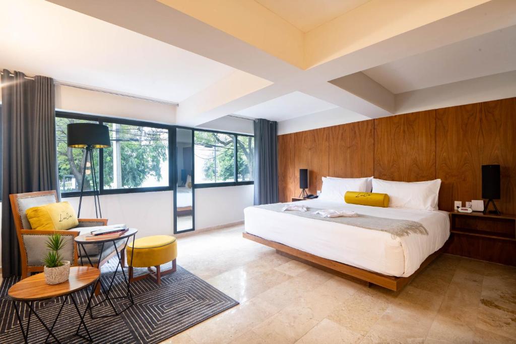 Modern hotel room with a large bed, wooden accent wall, two nightstands, a sitting area with chairs, small tables, a floor lamp, and large windows letting in natural light.