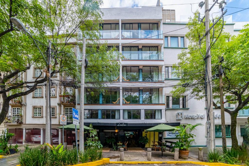 A modern white building with balconies and large windows, surrounded by trees. The ground floor has a café with outdoor seating and green umbrellas. Power lines and a yellow painted curb are visible in the foreground.