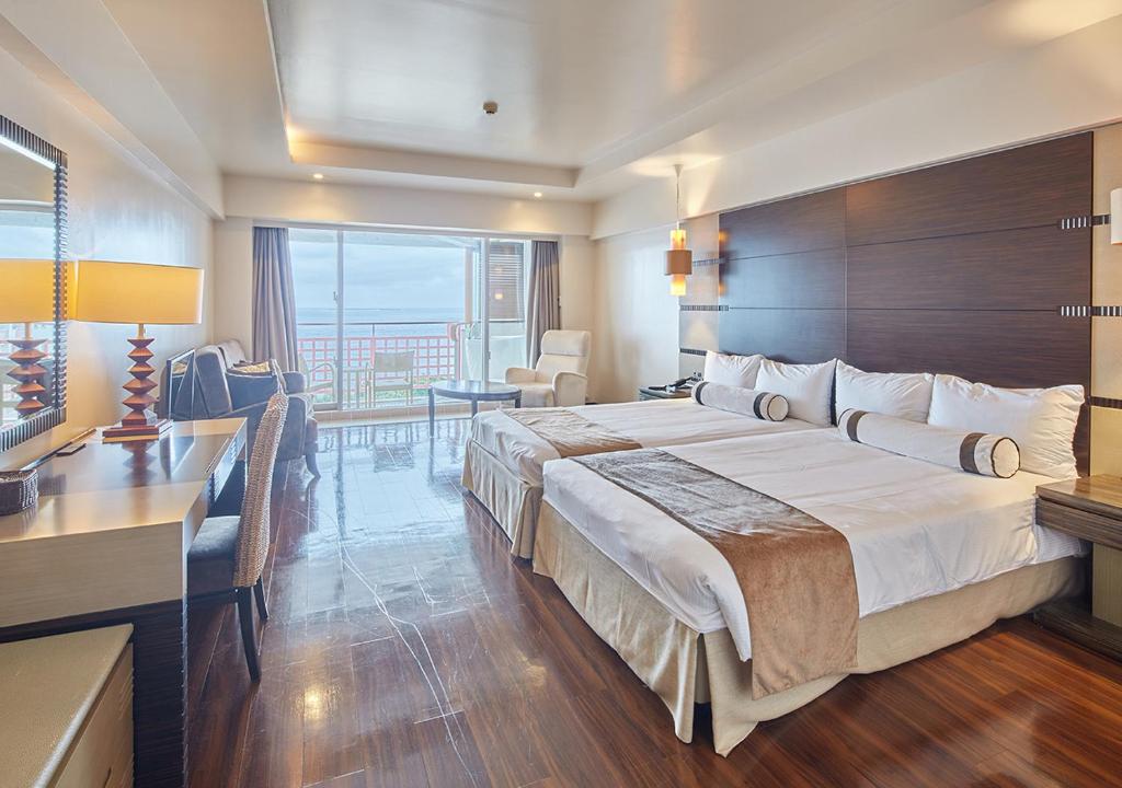 A modern hotel room with two large beds, a desk, a chair, warm lighting, and a sliding glass door opening to a balcony with a view of the sea. The room has wooden floors and neutral-colored decor.