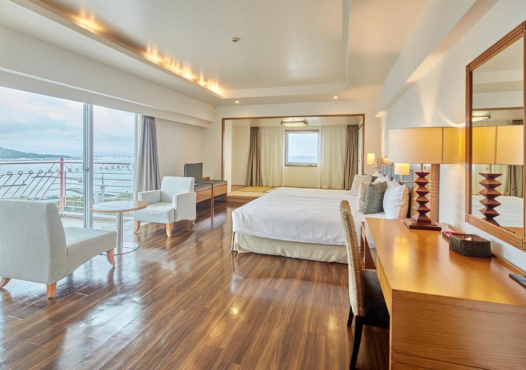 A spacious, modern hotel room with wooden flooring, a large bed, white chairs, a desk with lamps, and floor-to-ceiling windows offering a scenic view of water and distant hills.