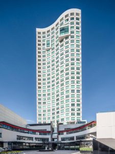 A tall, modern high-rise building with white and green exterior and curved top, stands against a clear blue sky above a lower curved structure in the foreground.