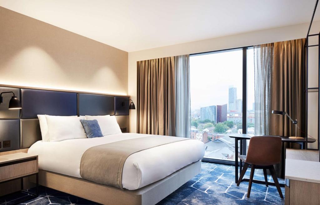 A modern hotel room with a large bed, nightstands, a desk with a chair, and a floor-to-ceiling window with curtains, offering a city view. The decor is neutral with blue and beige accents.