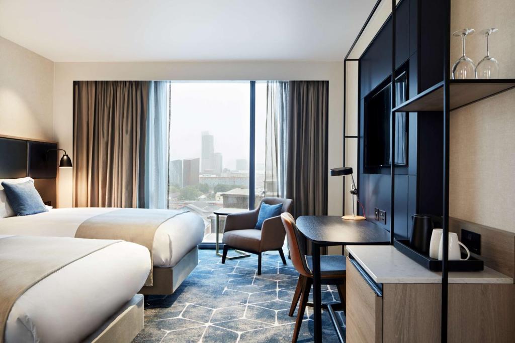 Modern hotel room with two double beds, a desk with a chair, and a large window offering a city skyline view. The room features neutral colors, patterned carpet, and contemporary furnishings.