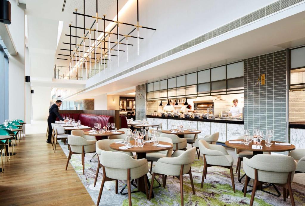 A modern restaurant with round wooden tables, beige chairs, and a patterned rug. Open kitchen in the background, chefs cooking, and a staff member setting a table. Large windows provide natural light.