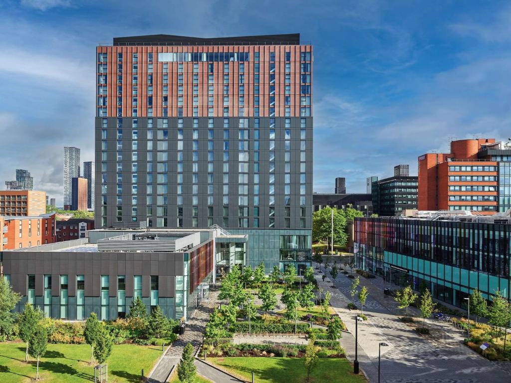 A modern multi-story building with a red and gray facade stands behind landscaped gardens with trees and paths, set against a blue sky and surrounded by other urban buildings.