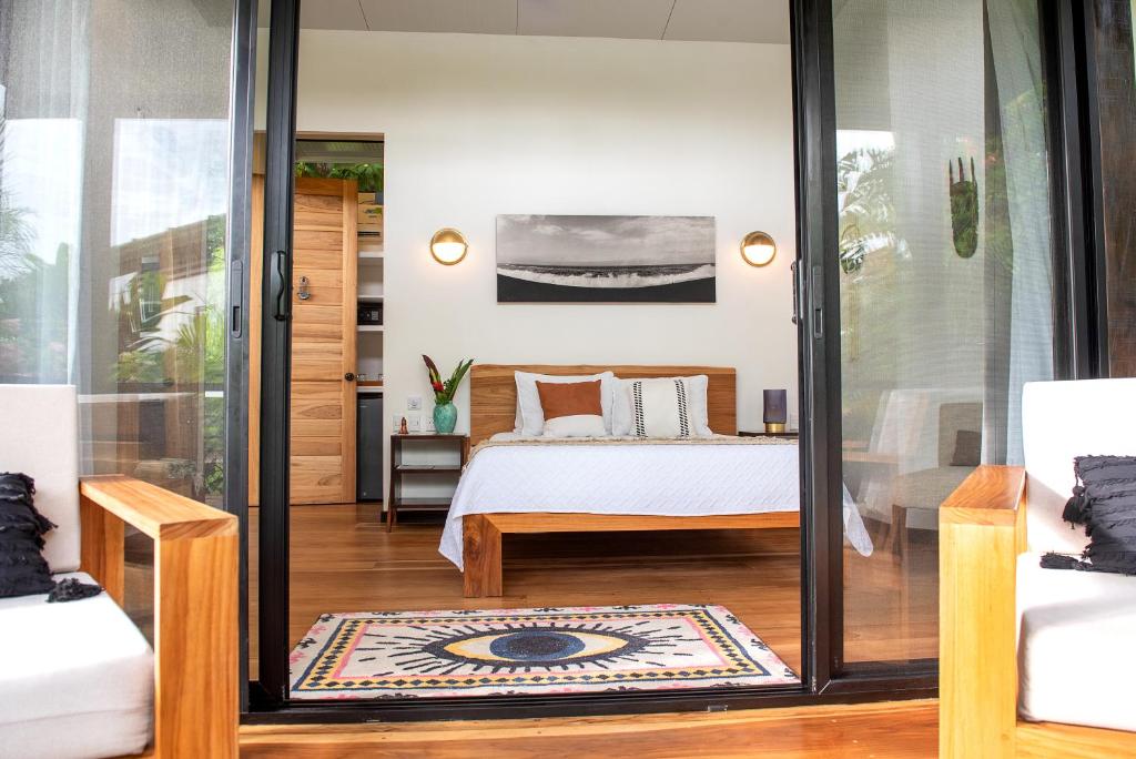 A cozy, modern bedroom with a wooden bed, white bedding, and a large monochrome photo above the headboard, viewed through open sliding glass doors. Two wooden chairs and a colorful rug are on the patio outside.