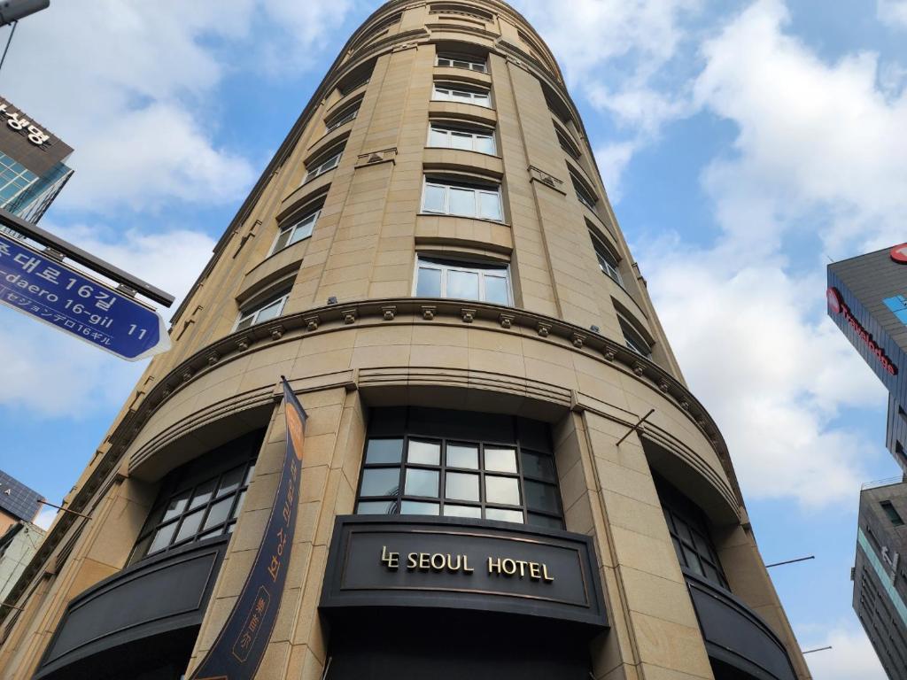 A tall, beige stone building with curved architecture and multiple windows is seen from below. A sign reads Le Seoul Hotel. Blue sky with clouds and street signs are visible.