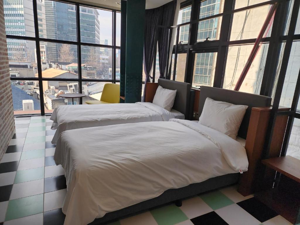 Two neatly made twin beds in a modern room with large floor-to-ceiling windows, checkerboard tile floor, yellow chair, and city buildings visible outside.
