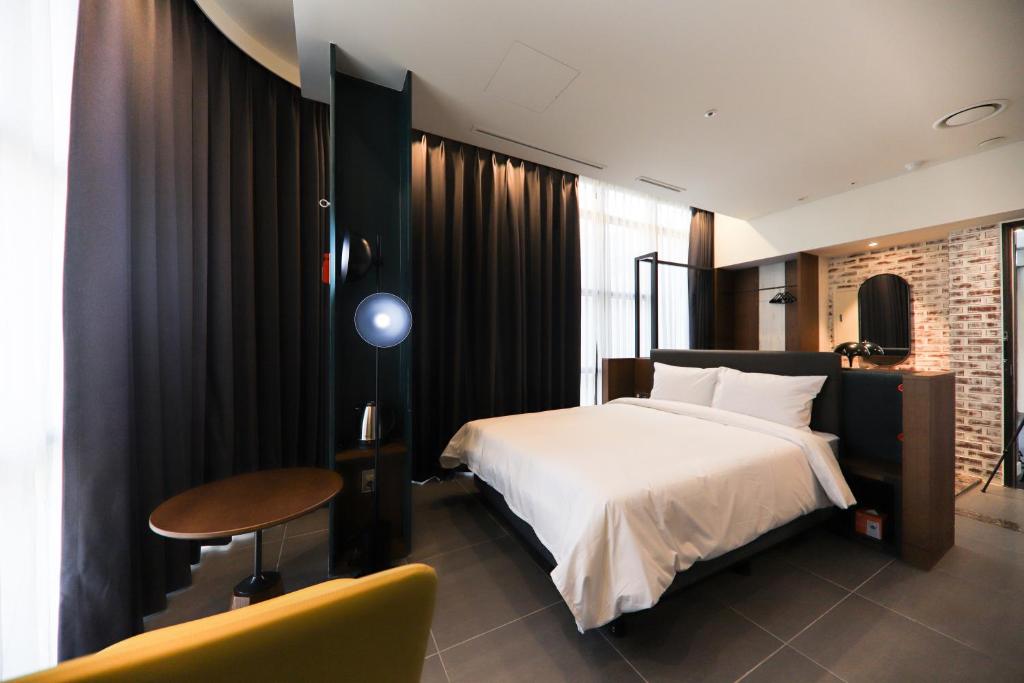A modern hotel room with a large bed, white linens, dark curtains, a round table, a yellow chair, and a sleek bathroom area with a brick accent wall visible in the background.