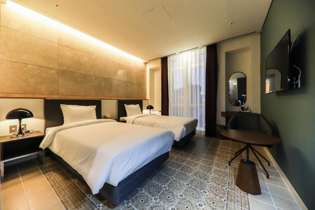 Modern hotel room with two single beds, white bedding, a TV on the wall, a small round table with a chair, patterned tile flooring, and large window with sheer curtains letting in natural light.