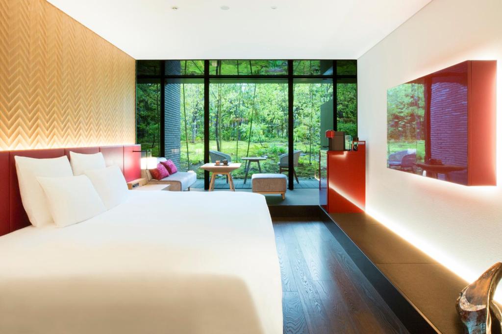 Modern hotel room with a large bed, wall-mounted TV, seating area, and floor-to-ceiling windows overlooking a lush green garden. The room has stylish décor with warm lighting and contemporary furnishings.