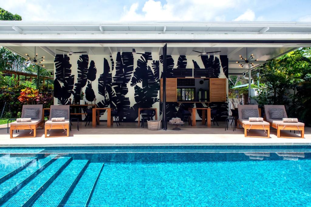 Outdoor pool with clear blue water in front of a covered patio featuring lounge chairs, tables, and a wall with bold black leaf patterns, surrounded by lush greenery under a bright sky.