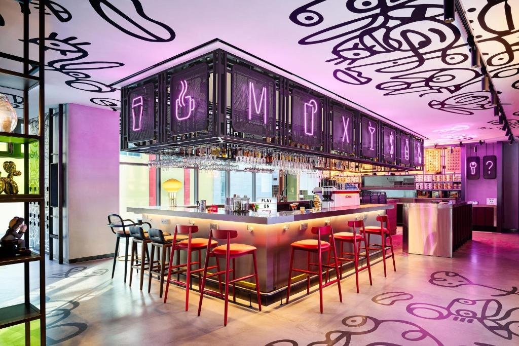 A modern bar with neon pink lighting, red barstools, and playful black line art on the ceiling and floor. The bar is surrounded by glass walls, and illuminated icons are displayed above the bar area.