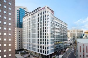 A modern, multi-story office building with large windows stands on a city street, surrounded by other tall buildings under a clear blue sky.