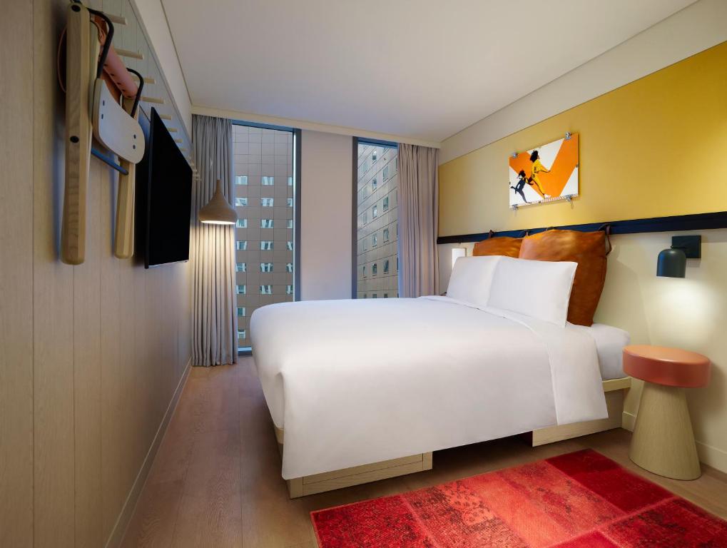 A modern hotel room with a large bed, white linens, brown leather headboard, a wall-mounted TV, floor-to-ceiling windows with city views, a red rug, and contemporary decor on a yellow accent wall.