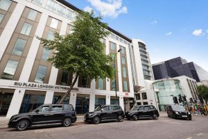 Four black London taxis are parked in front of the RIU Plaza London Victoria Hotel, a modern white and brown building with large windows and a tree by the entrance, under a blue sky with a few clouds.