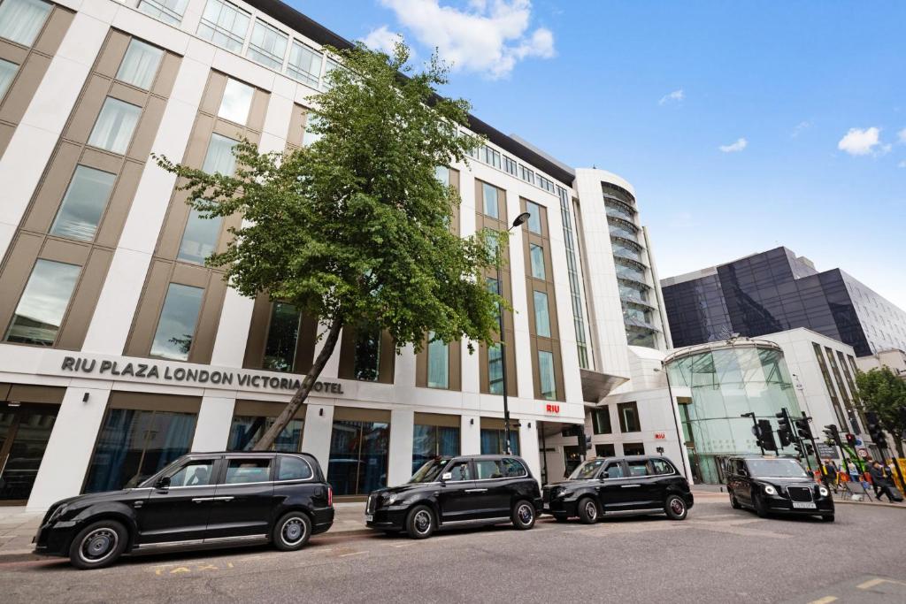 Four black London taxis are parked in front of the RIU Plaza London Victoria Hotel, a modern white and brown building with large windows and a tree by the entrance, under a blue sky with a few clouds.