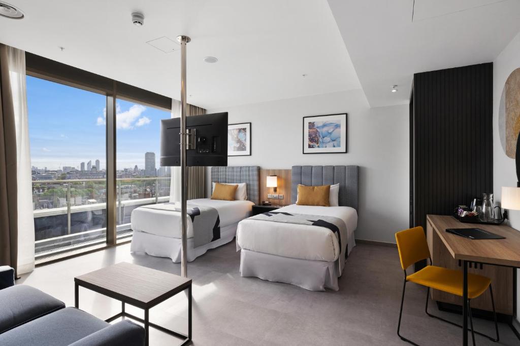 Modern hotel room with two single beds, a wall-mounted TV, a desk with a yellow chair, and a large window offering a city view and access to a balcony. The room features minimalist decor and neutral tones.