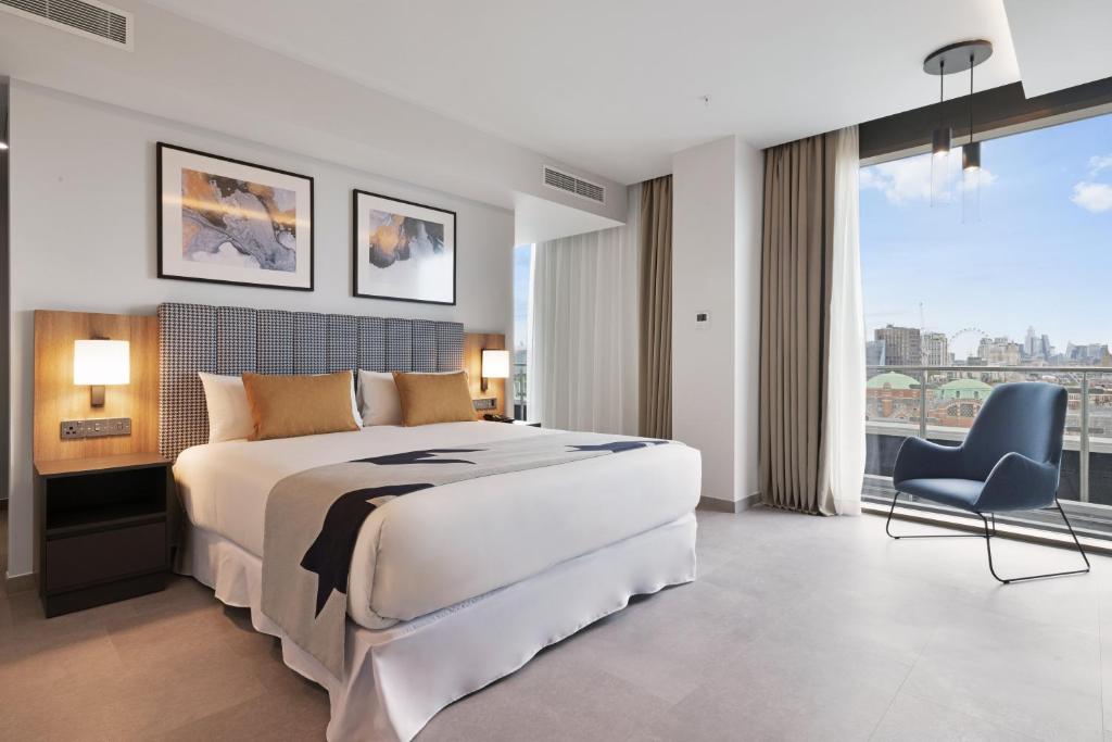 Modern hotel room with a large bed, two bedside tables with lamps, abstract art above the headboard, a chair by a floor-to-ceiling window, and a city view outside. The decor is minimalist and bright.